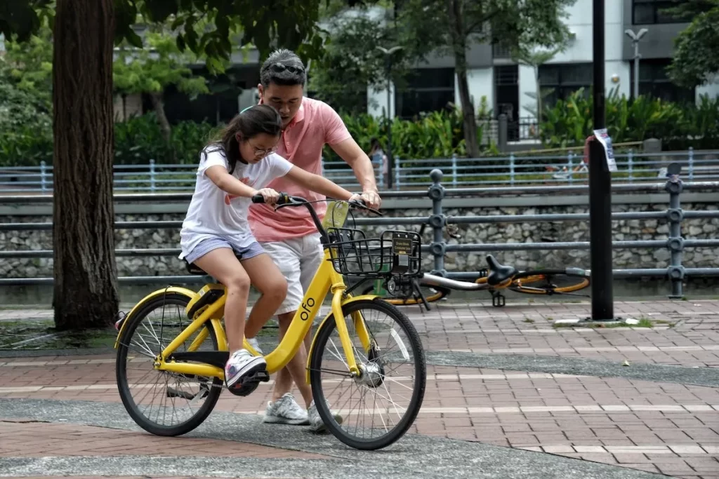 Um pai ajudando sua filha a pedalar uma bicicleta infantil amarela em um parque.