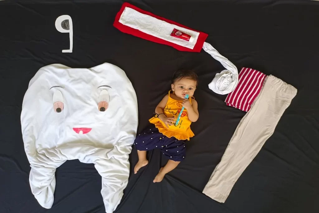 Bebê deitado em um lençol preto com um acessório de escova de dentes na mão, cercado por um dente gigante de tecido, um tubo de pasta de dente e roupas, com o número "9" indicando meses, em uma cena criativa de mêsversário focada no nascimento dos dentes.