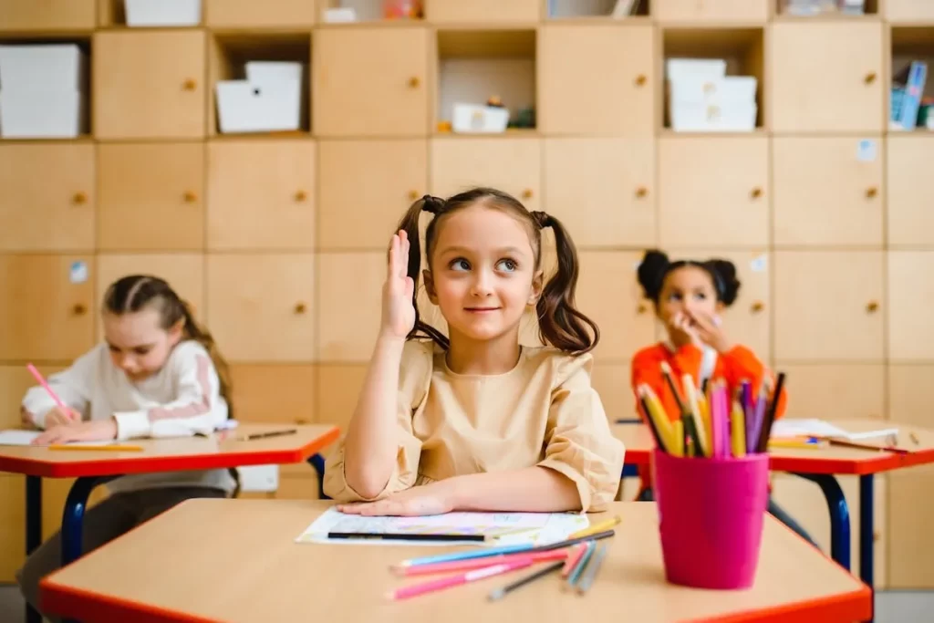Uma menina sorridente com tranças, levantando a mão na sala de aula, com outras crianças em segundo plano.