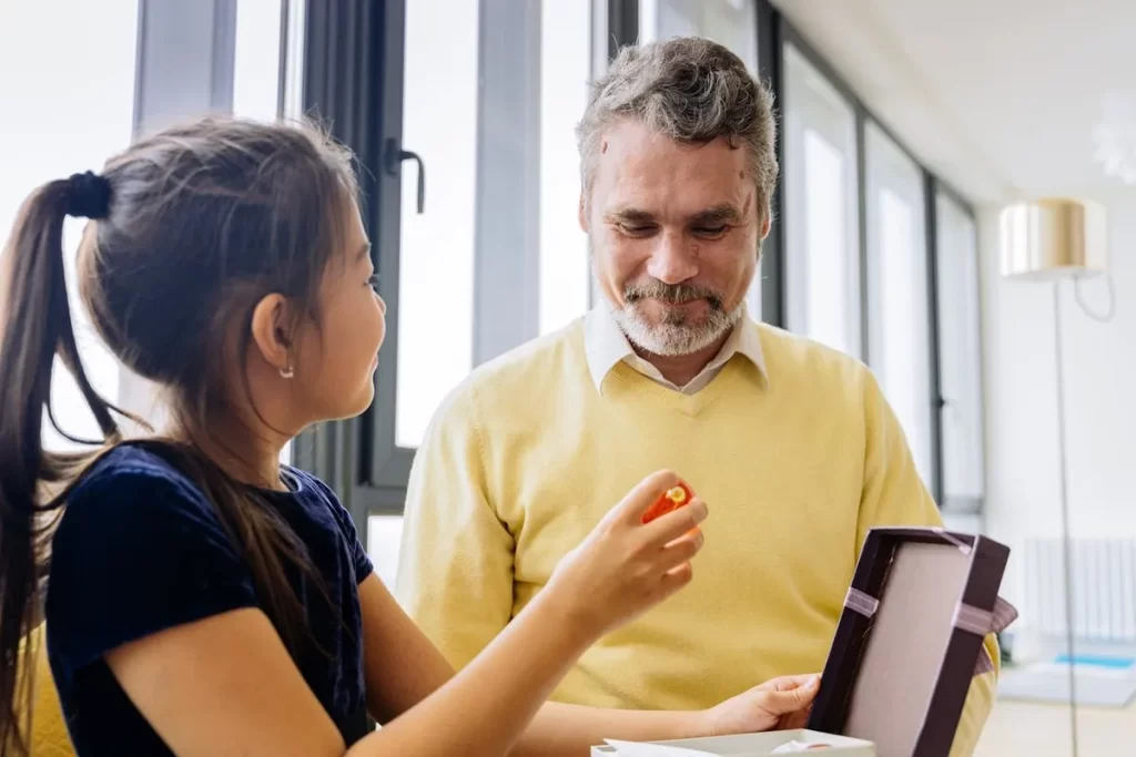 Menina sorrindo enquanto entrega um presente de Dia dos Pais para seu pai, que sorri olhando para o objeto.