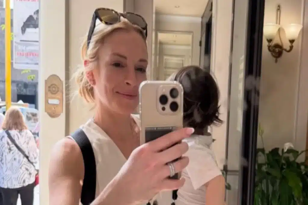 Lindsay Lohan, com os cabelos loiros presos e óculos de sol na cabeça, segura e tira uma selfie com seu filho, Luai, em um elevador ou ambiente interno.