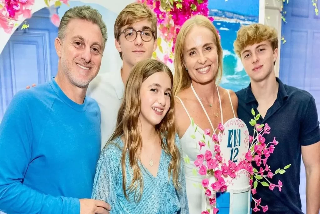 Família reunida, sorridente, posando para foto durante uma celebração especial, com decoração floral e bolo personalizado ao fundo.