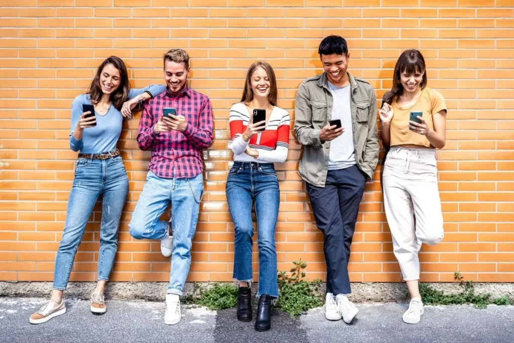 Grupo de jovens da Geração Z encostados em uma parede, sorrindo e interagindo com seus smartphones, representando conectividade e redes sociais.