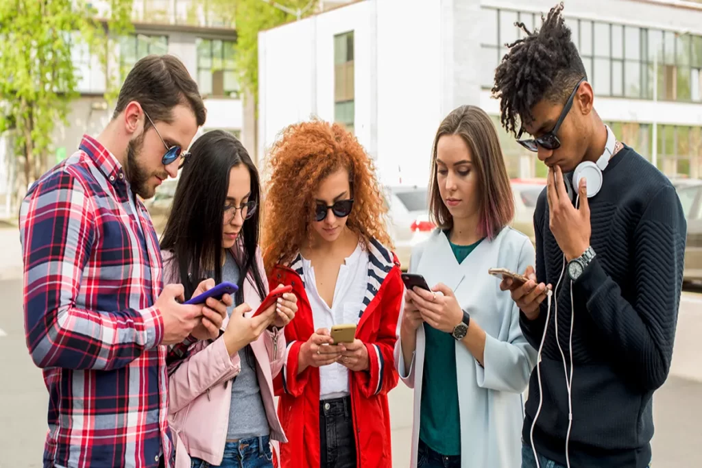 Grupo de jovens da Geração Z reunidos ao ar livre, todos concentrados em seus smartphones, representando o uso intenso de tecnologia e redes sociais.