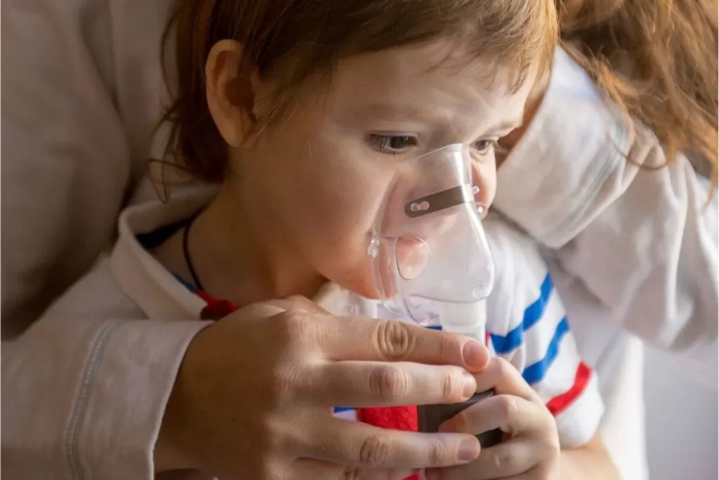 Criança pequena usando uma máscara de nebulização para tratar de um problema respiratório, representando uma doença como a coqueluche.