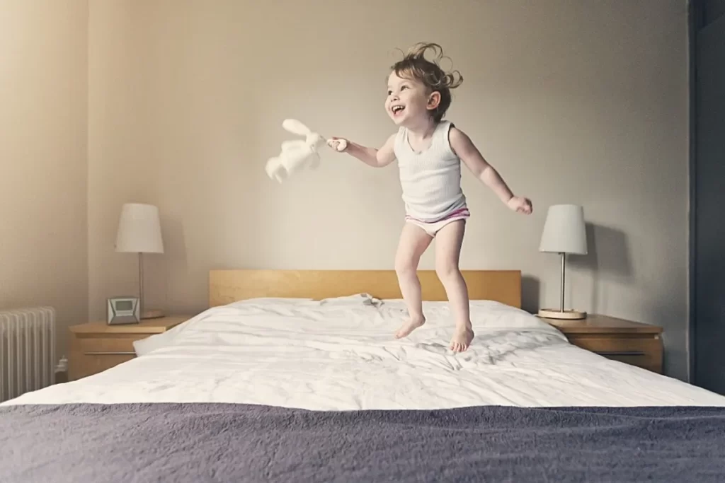 Criança pequena pulando na cama com um brinquedo de pelúcia, em uma cena que pode ser associada a hiperatividade.