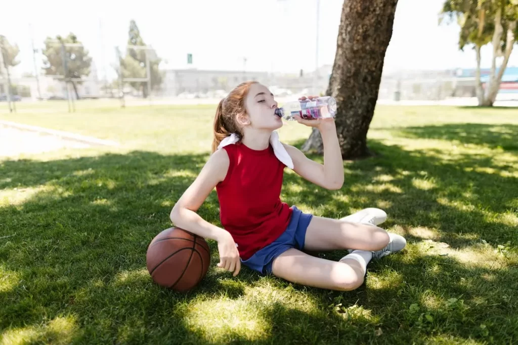 Menina com roupa esportiva sentada na grama, com uma bola de basquete ao lado, bebendo água de uma garrafa. A imagem levanta a questão se crianças podem tomar Gatorade para hidratação.