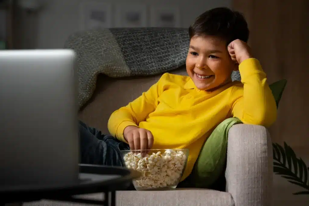 Criança sorridente assistindo desenho infantil no YouTube em um notebook, comendo pipoca.