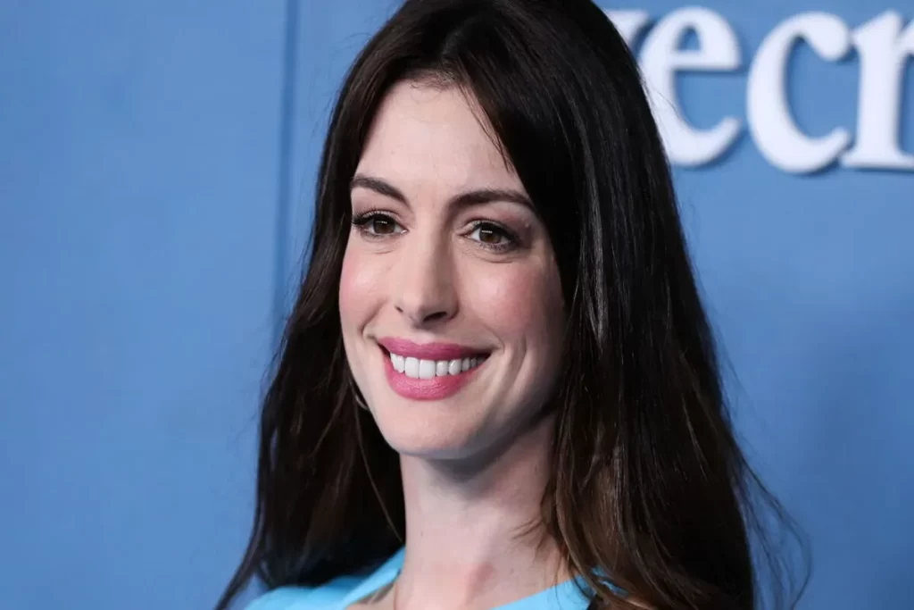 Close-up da atriz Anne Hathaway sorrindo, com maquiagem suave e cabelo solto, em frente a um fundo azul. A imagem é associada à busca "Anne Hathaway filhos".