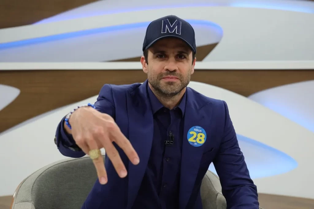 O empresário e coach Pablo Marçal, com um boné azul e um broche com o número 28, sentado em um estúdio de TV, gesticulando com a mão direita.