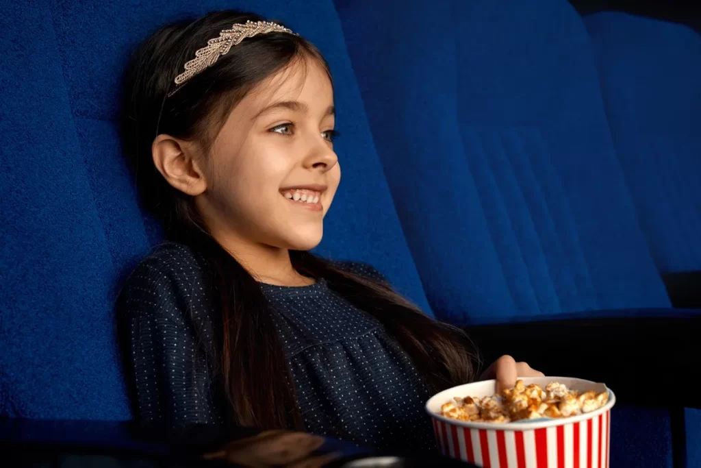 Garotinha sorridente com tiara brilhante sentada em poltrona azul de cinema segurando balde de pipoca