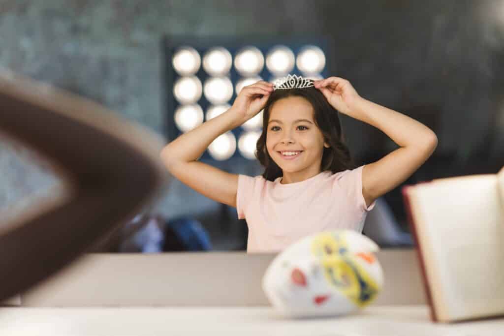 Menina sorridente coloca uma coroa na cabeça enquanto se olha no espelho em um camarim iluminado.
