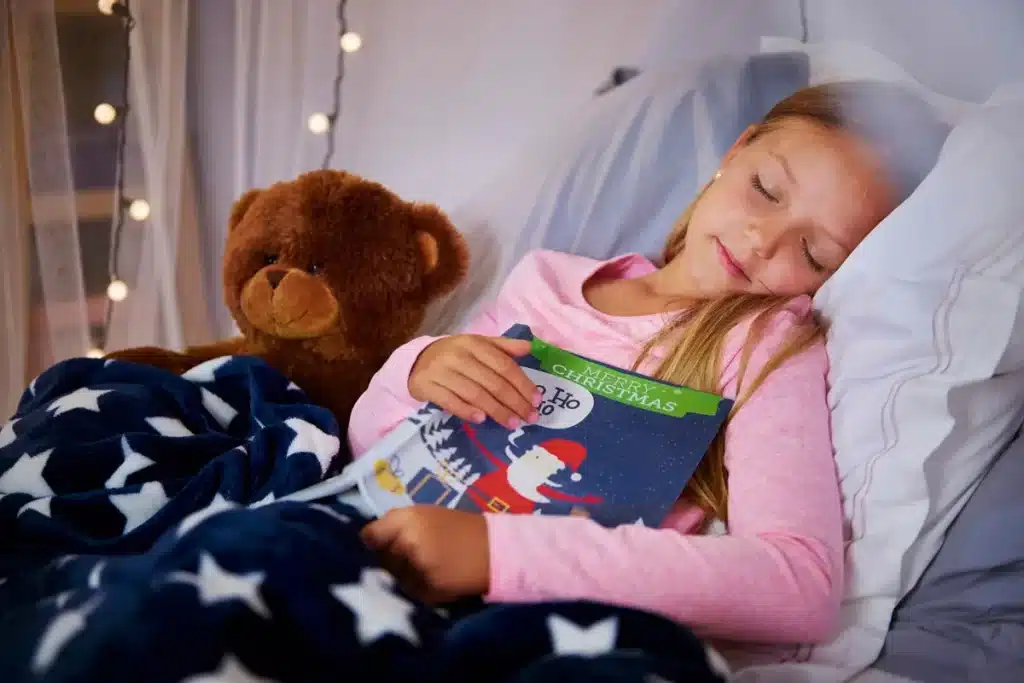 Menina loira dormindo com um livro de Natal nas mãos, deitada na cama ao lado de um urso de pelúcia e coberta com manta azul de estrelas