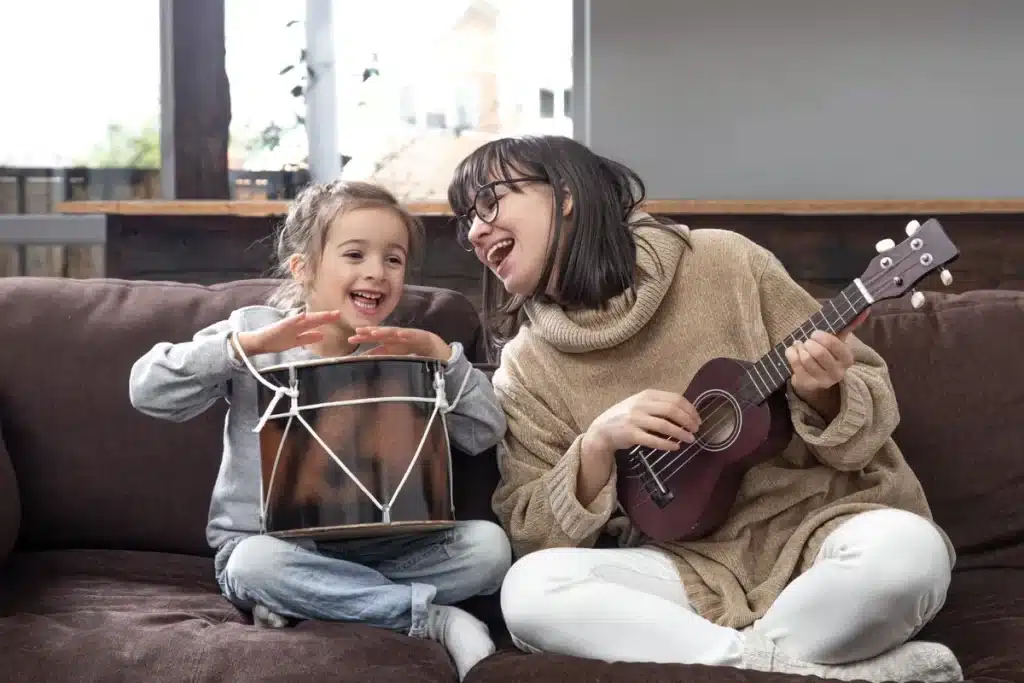 Uma mulher adulta e uma criança sentadas em um sofá marrom, tocando instrumentos musicais (a mulher toca um ukulele e a criança bate em um pequeno tambor) com sorrisos de alegria, representando uma sessão de musicoterapia infantil ou familiar.