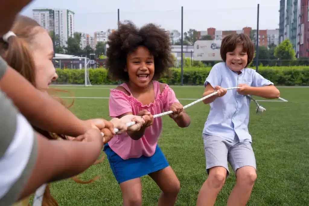 Crianças rindo e brincando de cabo de guerra em um campo de grama, com prédios ao fundo.