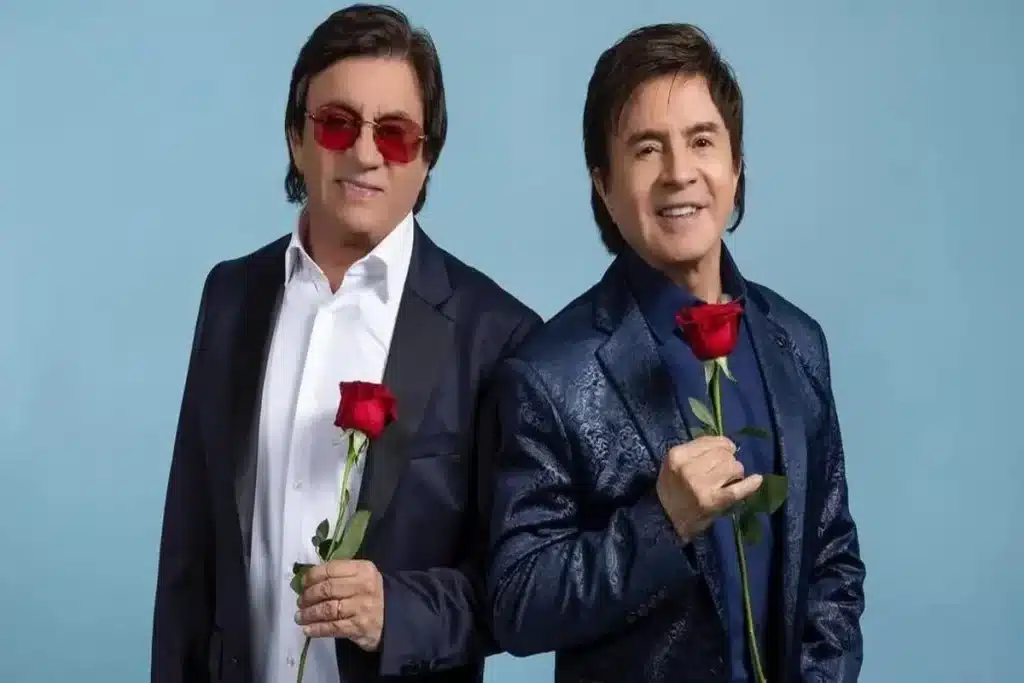 Chitãozinho e Xororó, a famosa dupla sertaneja brasileira, posando lado a lado com um sorriso e cada um segurando uma rosa vermelha, contra um fundo azul claro.