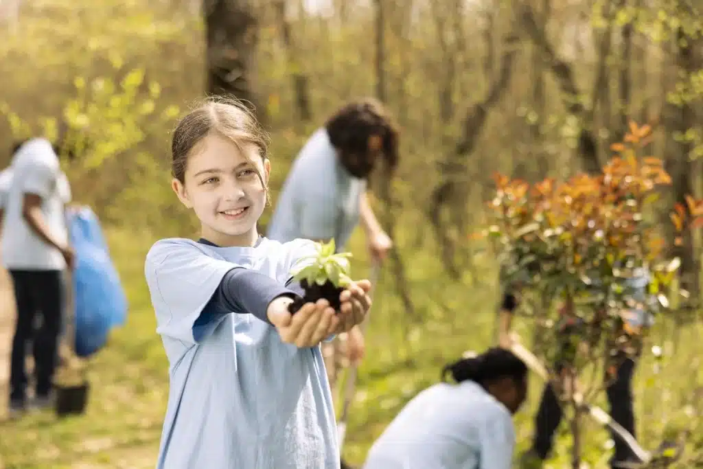 Menina sorridente segurando uma muda de planta em um ambiente ao ar livre, cercada por outras crianças participando de atividades de plantio.