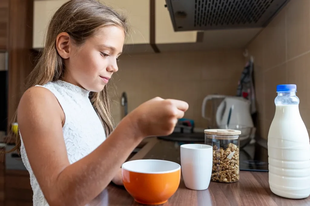 Menina preparando café da manhã com leite e cereal na cozinha, representando alimentação infantil e suplementação nutricional.
