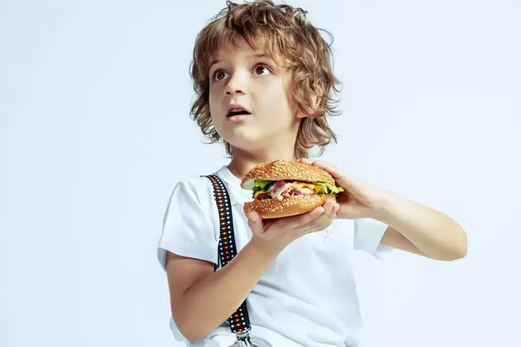  Menino com cabelo cacheado segurando um hambúrguer grande com expressão surpresa e olhar voltado para cima, vestindo camiseta branca e suspensórios coloridos.