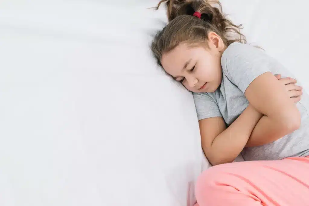 Menina deitada segurando o abdômen com expressão de desconforto, vestida com roupa confortável, em um ambiente claro