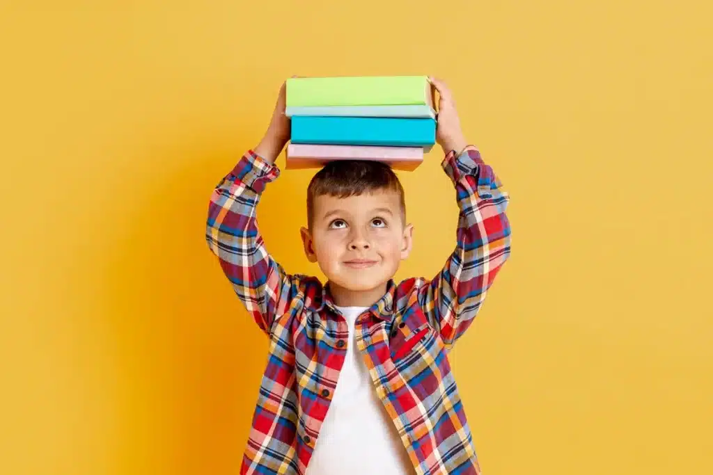 Menino sorridente segurando uma pilha de livros coloridos sobre a cabeça, com fundo amarelo vibrante