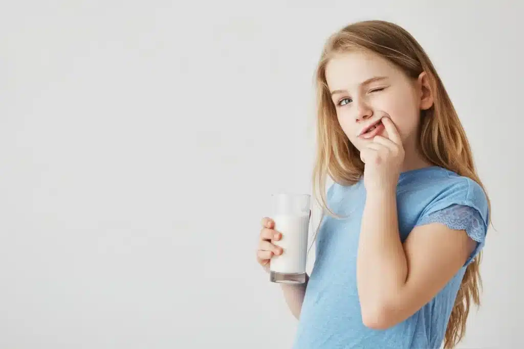 Menina com cabelos longos loiros, vestindo uma camiseta azul, segurando um copo de leite enquanto faz uma expressão divertida com um sorriso e pisca um olho.