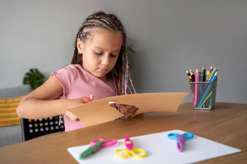 Menina concentrada cortando papel com tesoura colorida em uma mesa, cercada por lápis de cor e materiais de artesanato