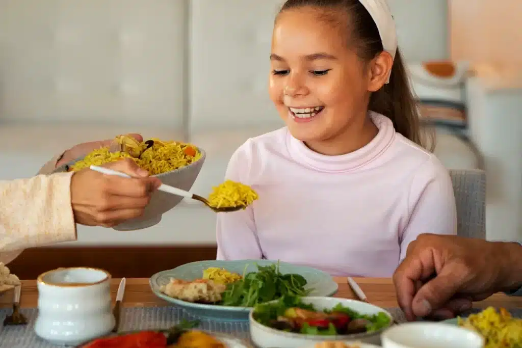 Menina sorridente de cabelo castanho e faixa branca recebe uma porção de arroz amarelo em seu prato durante uma refeição em família. A mesa está posta com pratos contendo saladas verdes, vegetais e frango grelhado, criando um ambiente acolhedor e nutritivo.