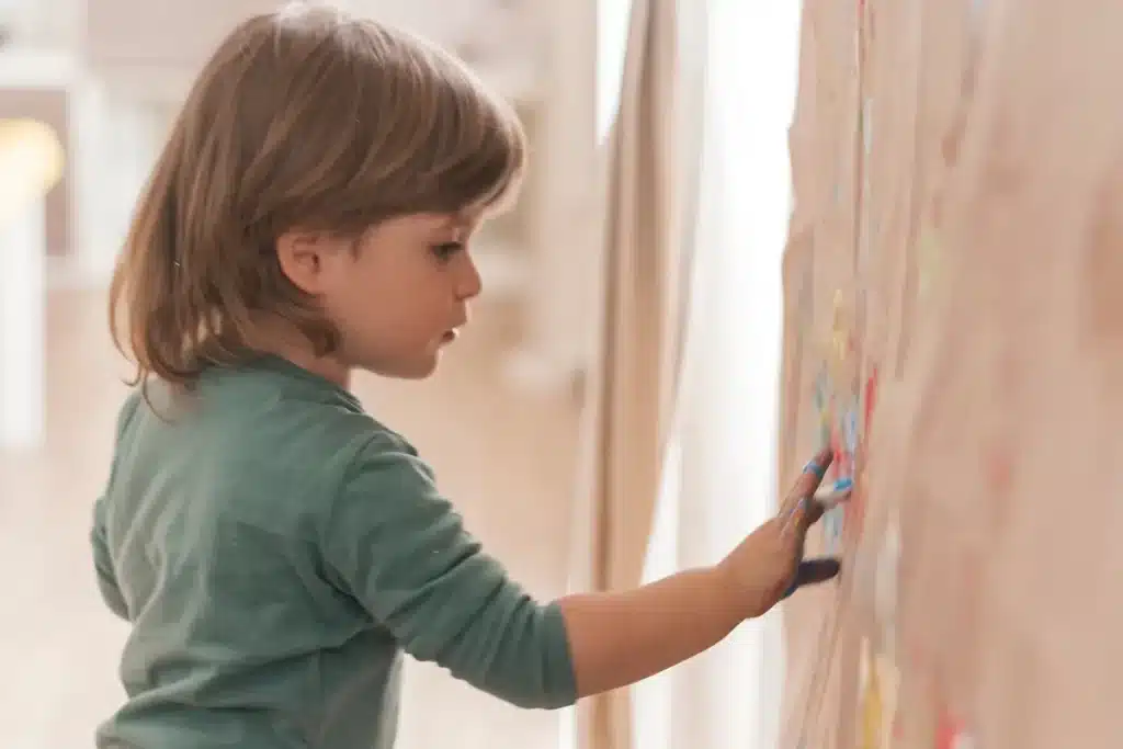 Criança pequena desenhando em um grande painel de papel com as mãos coloridas, participando de uma atividade criativa.