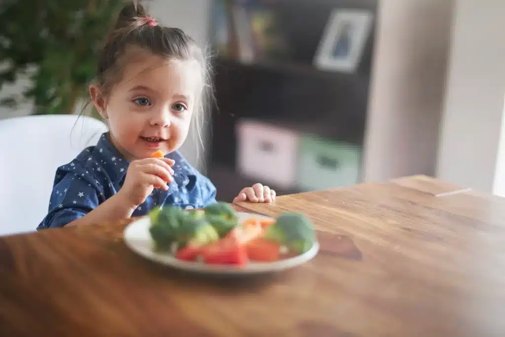 Menina sorridente com cabelo preso em um coque e blusa azul com estrelas sentada à mesa, segurando um pedaço de cenoura enquanto se alimenta. Na sua frente, um prato com brócolis, cenoura e tomate, representando uma refeição saudável e equilibrada.