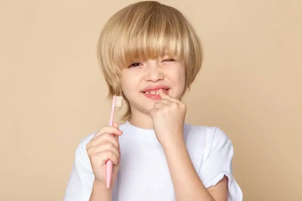 Menino loiro com camiseta branca segura uma escova de dentes rosa enquanto toca um dente de leite mole, simbolizando crescimento infantil e troca dentária.