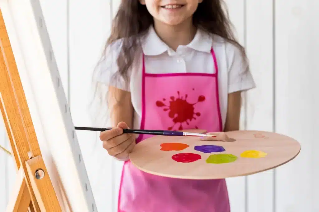 Menina segurando paleta de tinta colorida e pincel enquanto pinta em uma tela sobre um cavalete.