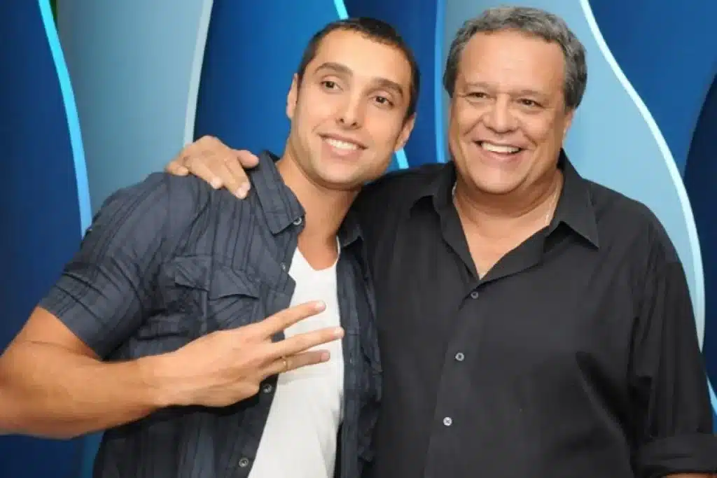 Dois homens sorridentes posam juntos em um evento, um vestindo camisa preta e o outro camisa azul com camiseta branca por baixo, contra um fundo azul estilizado.