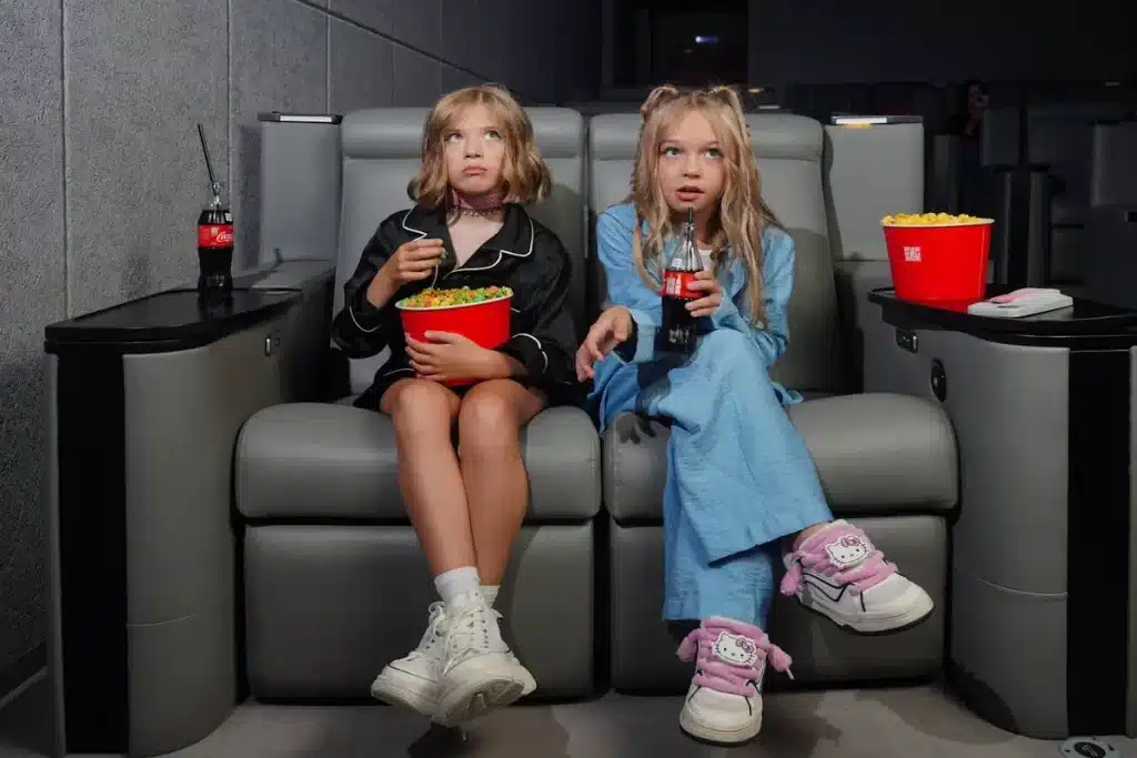 Duas crianças (meninas) sentadas em poltronas de luxo no cinema, assistindo a um filme infantil. Elas seguram pipoca e refrigerante e parecem concentradas ou surpresas com o que veem na tela.