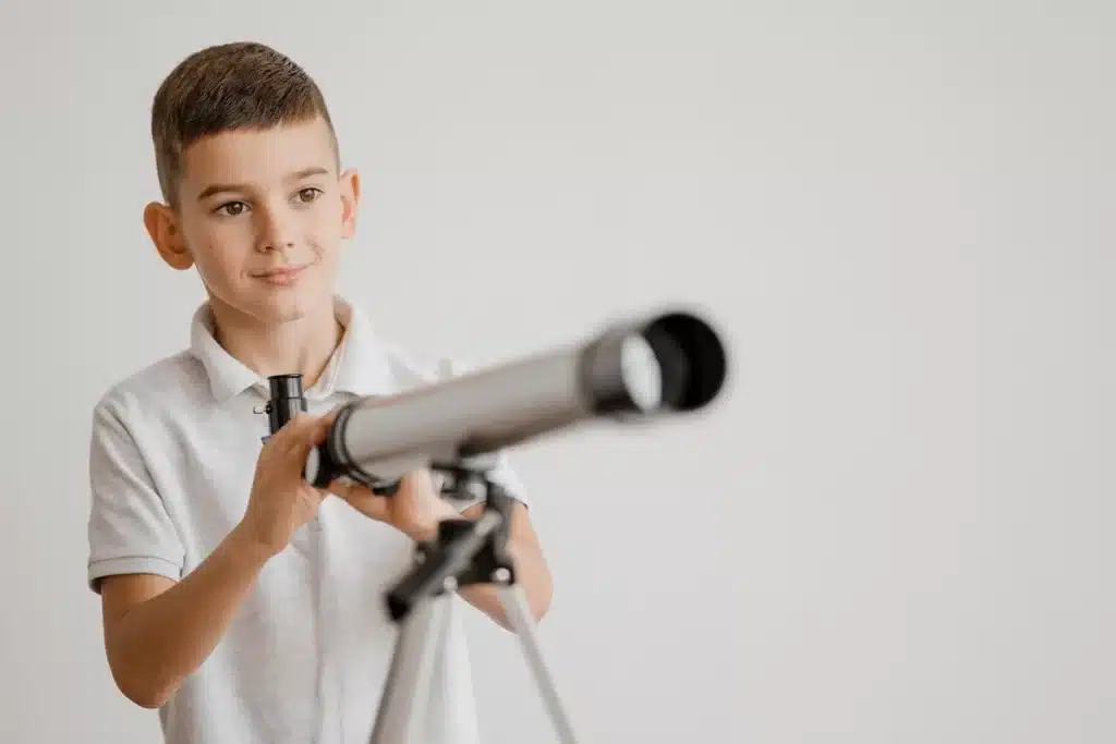 Menino sorridente com camiseta polo branca observando através de um telescópio em ambiente interno, representando brinquedo educativo para crianças.