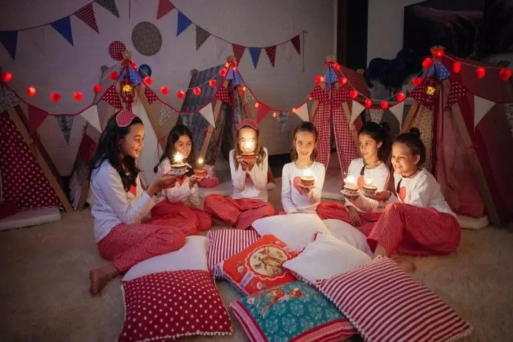 Crianças em uma festa do pijama dentro de cabaninhas decoradas, segurando cupcakes iluminados com velas.