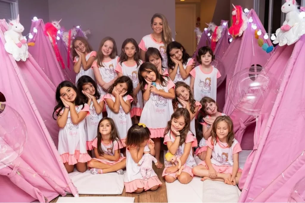 Grupo de meninas em uma festa do pijama com tema unicórnio, usando camisolas personalizadas e brincando em cabaninhas decoradas cor-de-rosa.