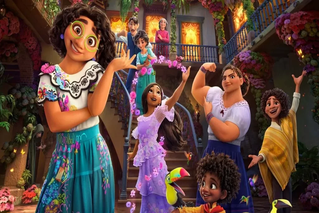 Cena colorida e vibrante do filme de animação Encanto da Disney, mostrando a Família Madrigal reunida na frente de sua casa mágica (Casita). A personagem principal, Mirabel (com óculos verdes e saia azul-turquesa), está em primeiro plano, com outros membros da família — como Luisa (a fortona, exibindo os músculos), Isabela (de vestido lilás) e Antonio (com o tucano e colete amarelo) — sorrindo e cantando em uma escadaria florida.