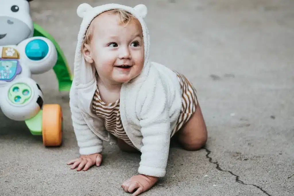 Bebê sorridente engatinhando no chão com moletom de orelhinhas e body listrado, ao lado de brinquedo educativo com rodinhas.