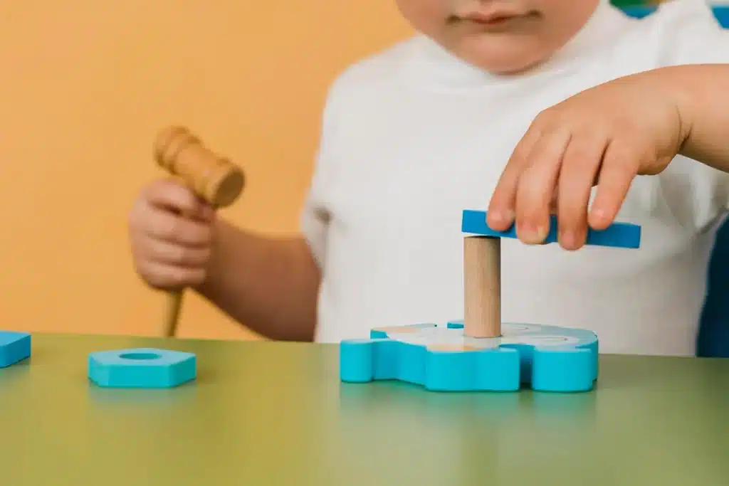 Criança brincando com brinquedo educativo, colocando peça azul sobre um pino de madeira com martelo de brinquedo ao lado