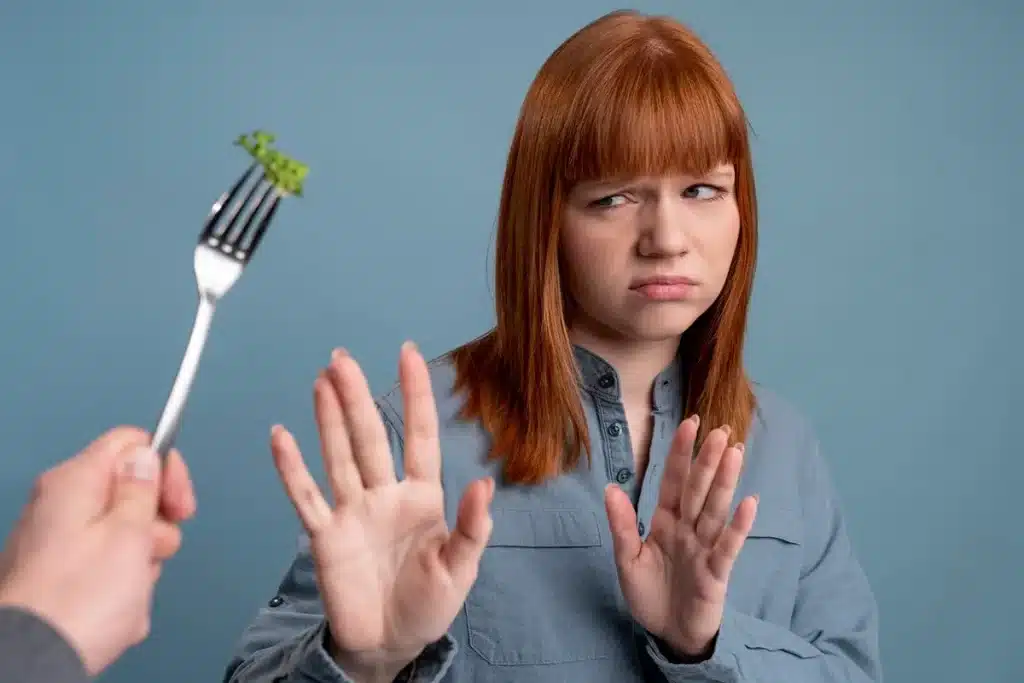 Mulher com cabelo vermelho e camisa azul claro, mostrando expressão de desgosto enquanto recusa um pedaço de vegetais com as mãos levantadas, como sinal de rejeição, com fundo azul.