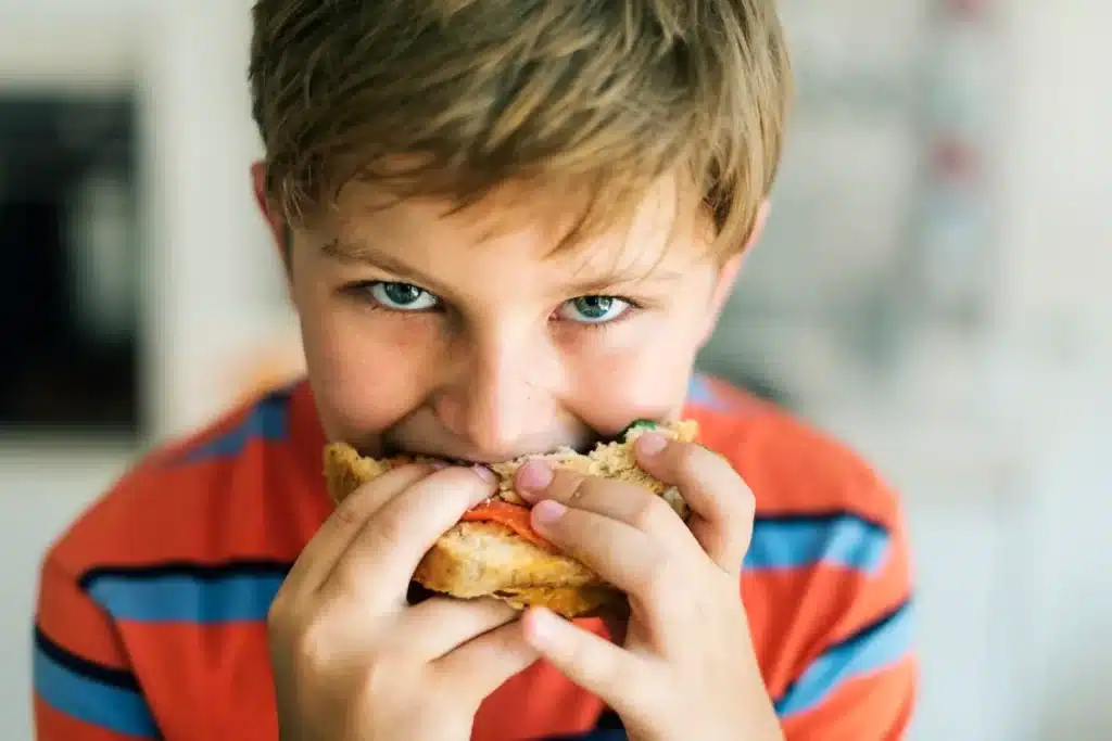 Criança comendo um sanduíche, segurando com as mãos enquanto olha para a câmera com expressão satisfeita.