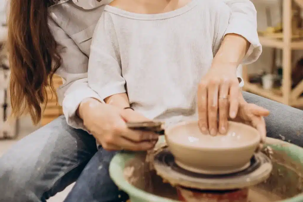 Imagem de uma criança e um adulto moldando argila em uma roda de oleiro, criando uma peça de cerâmica. As mãos de ambos guiam a argila em um processo criativo.