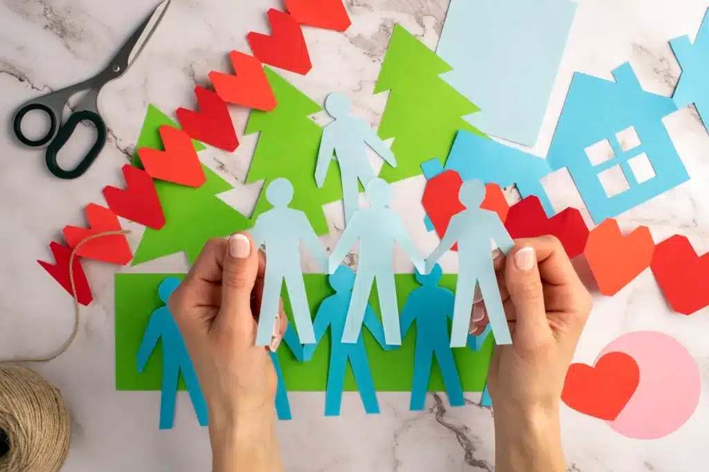 Mãos segurando figuras de papel representando membros de uma família, com recortes coloridos de corações, árvores e casas ao fundo.
