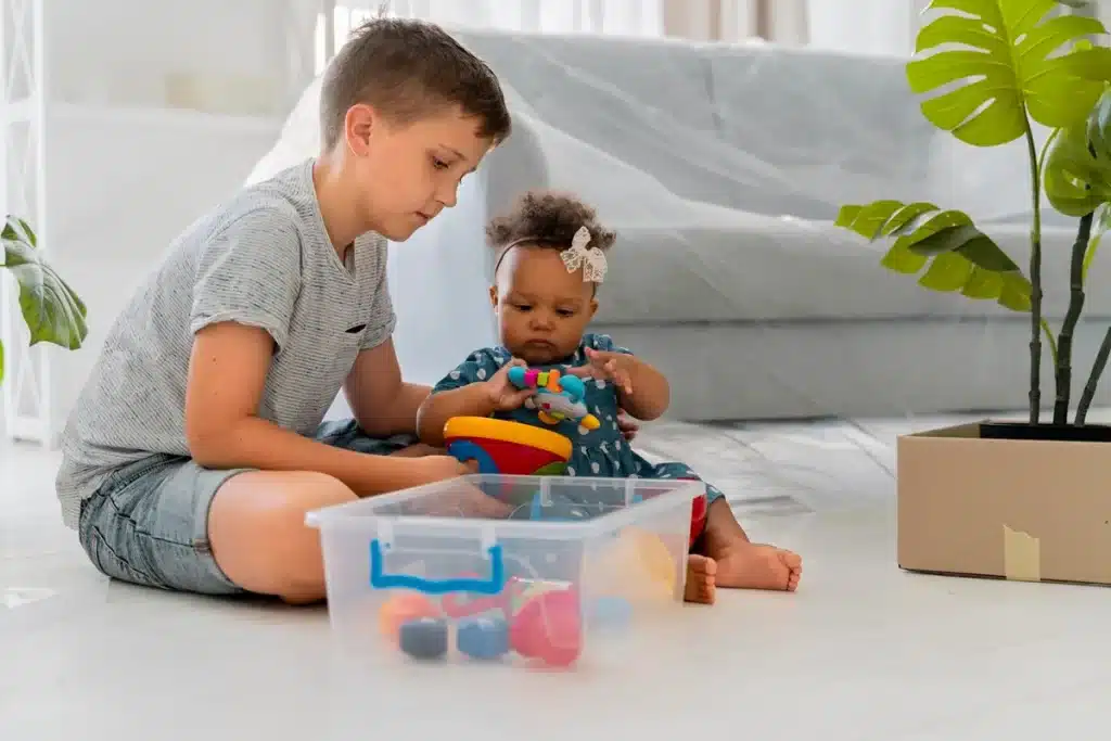 Criança mais velha ajudando bebê a brincar com brinquedos coloridos em uma caixa sensorial.