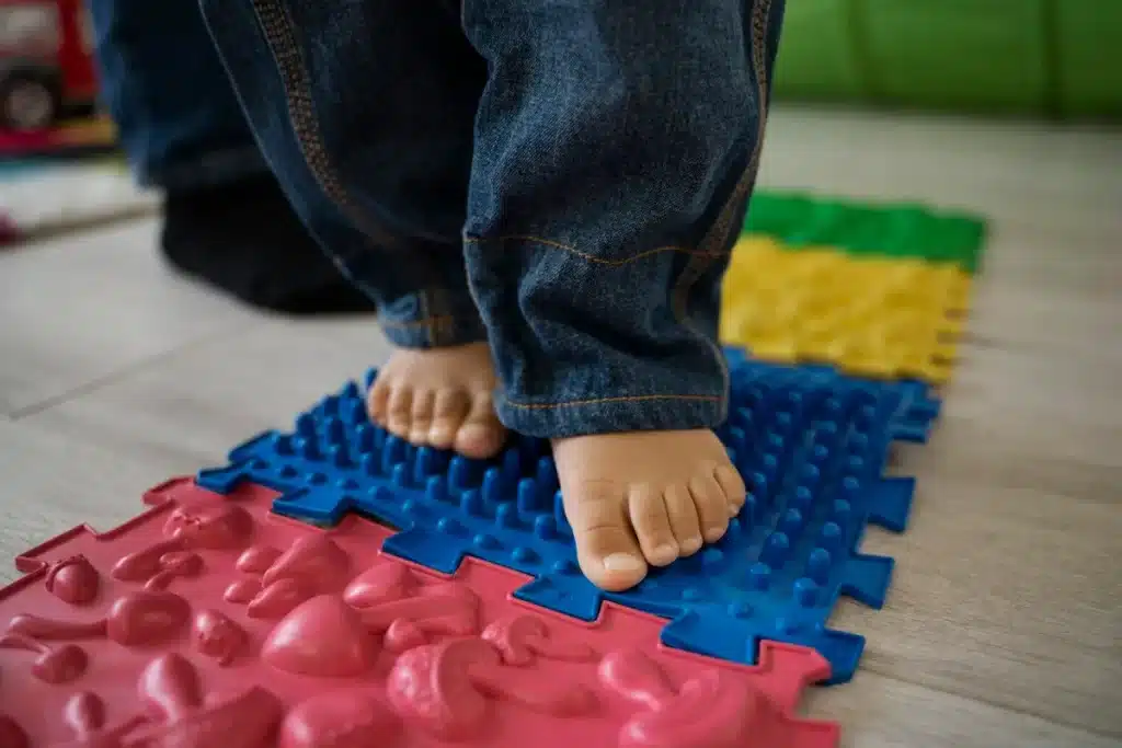 Criança caminhando descalça sobre tapetes sensoriais coloridos, com diferentes texturas para estimulação do circuito motor.