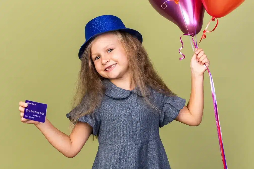 Criança com chapéu de festa segurando um cartão de crédito e balões coloridos, sorrindo em um fundo verde