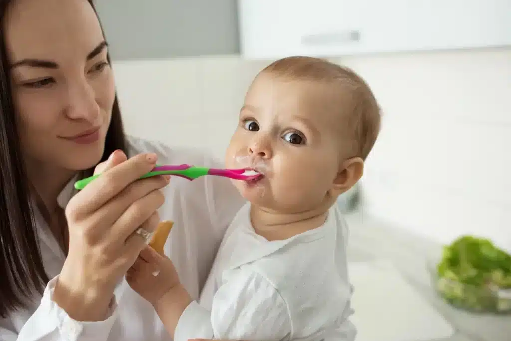 Mãe segurando escova de dentes com pasta e alimentando bebê, que está segurando um biscoito enquanto olha para a câmera. O cenário é uma cozinha clara e moderna.
