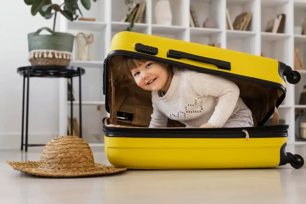 Menino pequeno sorrindo dentro de mala de viagem amarela aberta em ambiente doméstico, com chapéu de palha ao lado.
