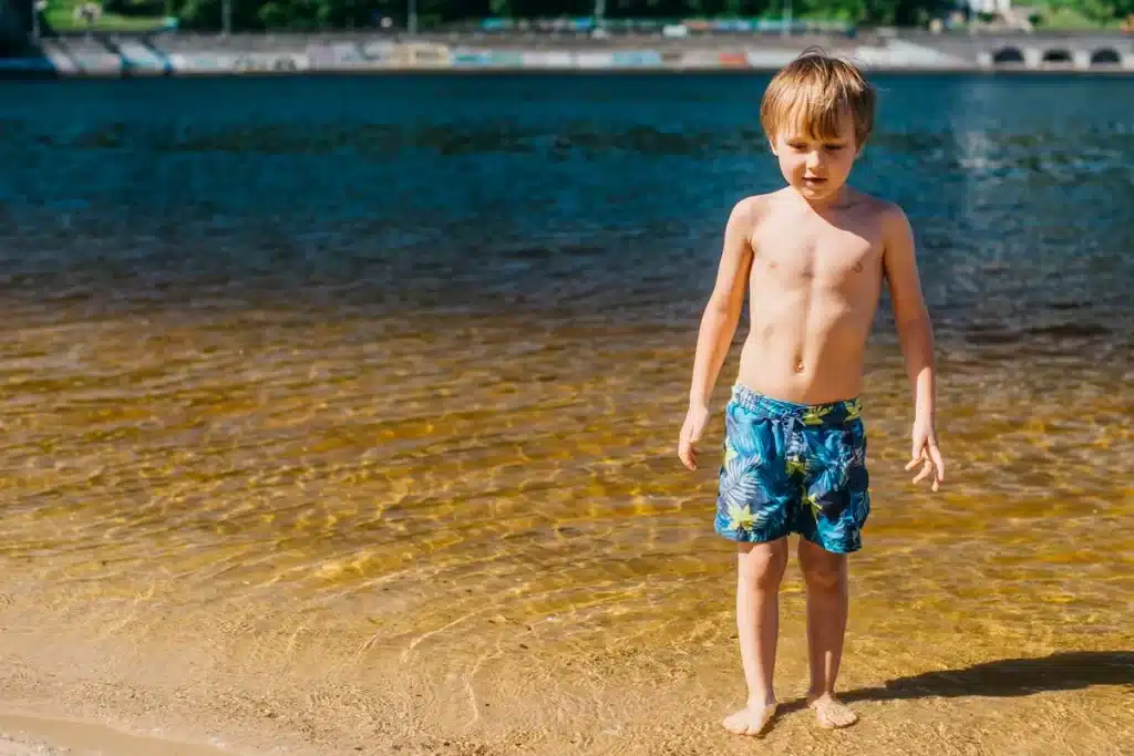 Criança com shorts de praia azul e estampado, brincando na água rasa de praia. Ele está descalço e sorrindo enquanto explora a água, com o cenário de uma praia ao fundo